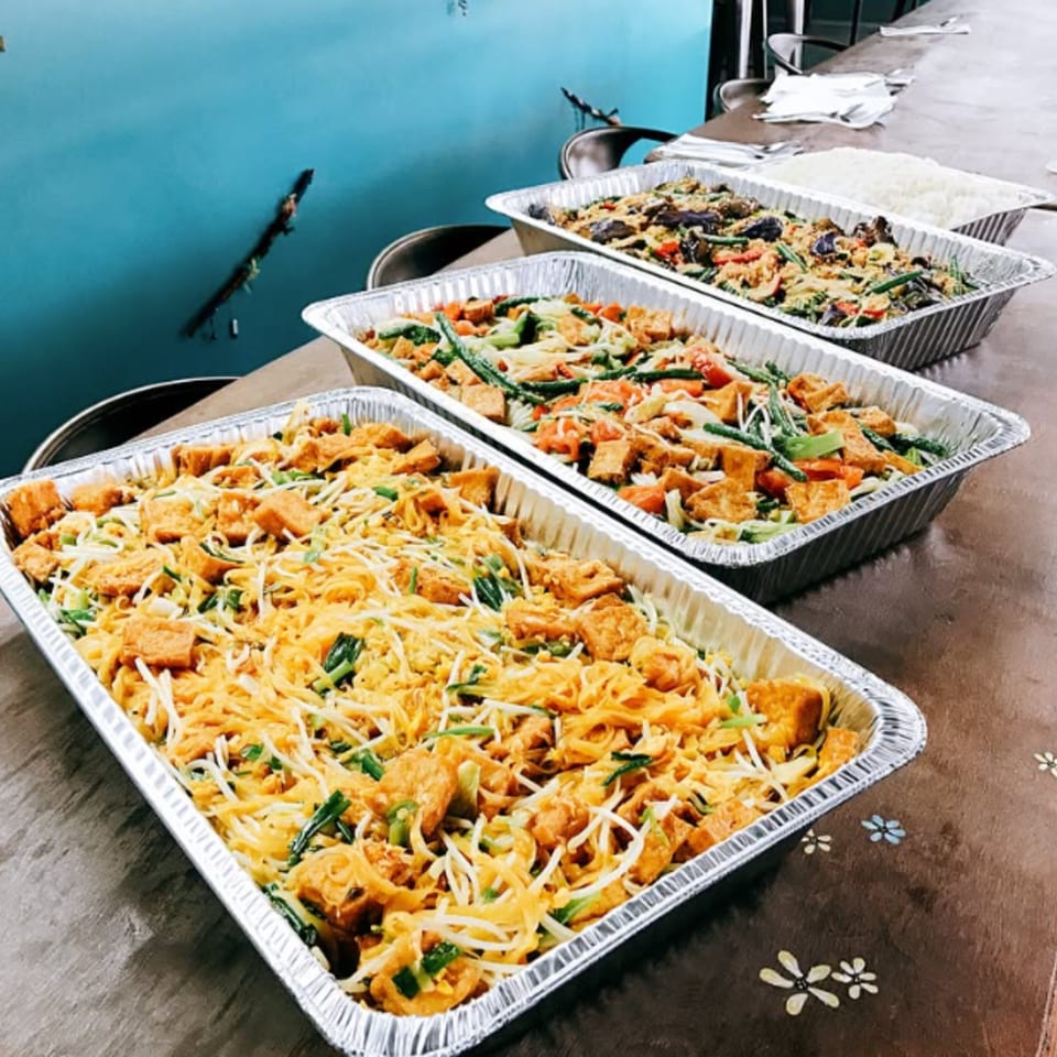 Three foil trays of Asian cuisine, including noodles with tofu, vegetables, and rice, on a table.