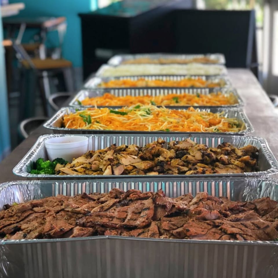 Multiple catering trays of Asian cuisine, including noodles, grilled chicken, and sliced beef.