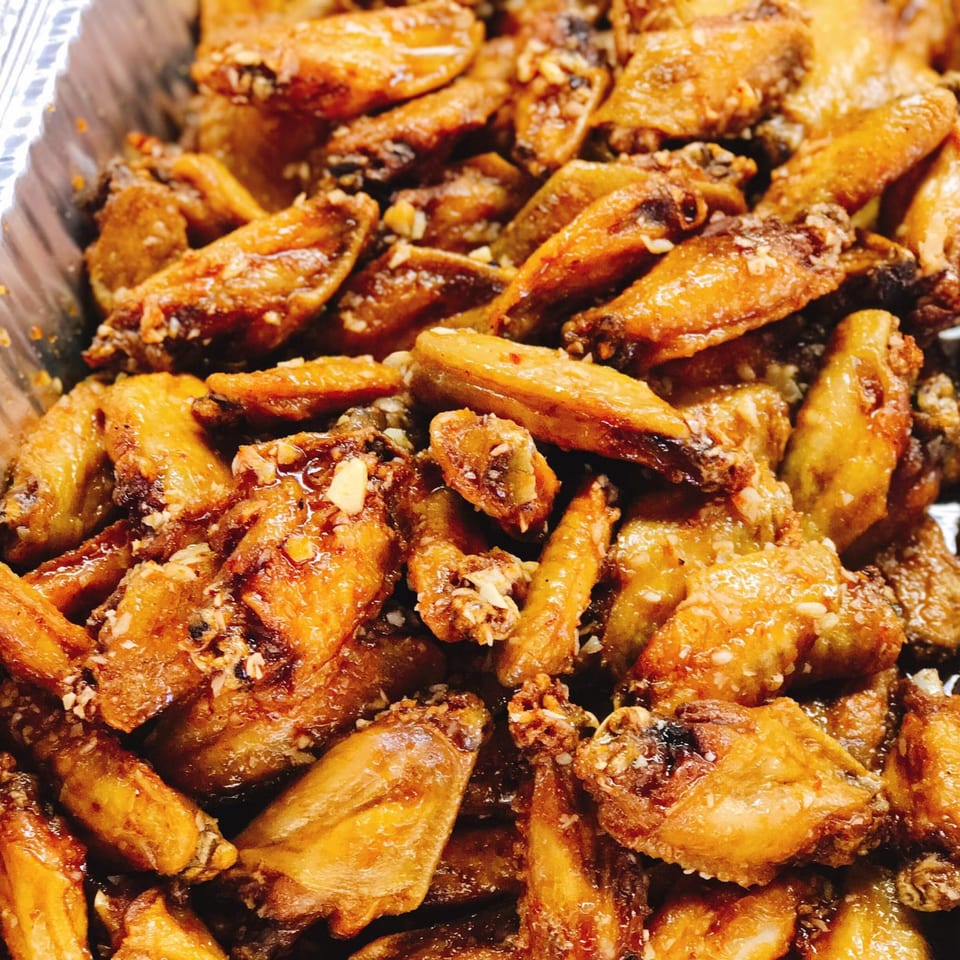 Close-up of golden-brown Asian chicken wings, glazed and sprinkled with chopped nuts in a foil tray.